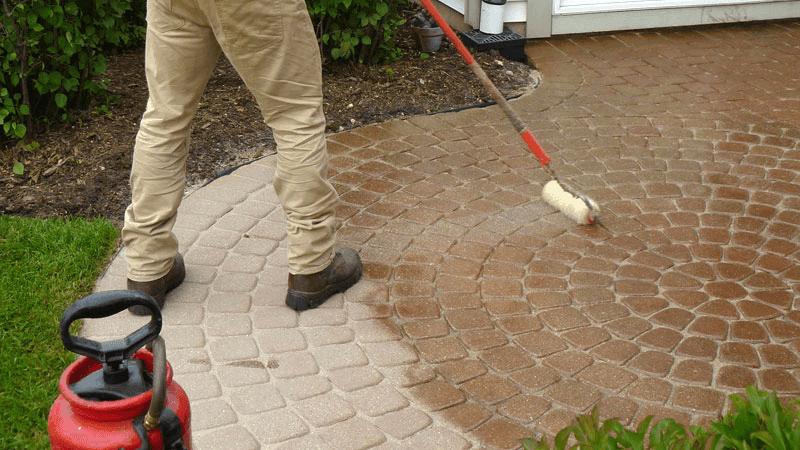 Person applying sealant to cobblestone patio with a roller, red spray bottle nearby.